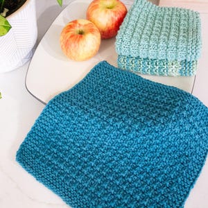 May include: A set of three square crochet washcloths featuring a textured waffle stitch pattern. One washcloth is a deep teal blue, while the other two are a lighter sage green. The cloths are arranged with two red and yellow apples nearby.