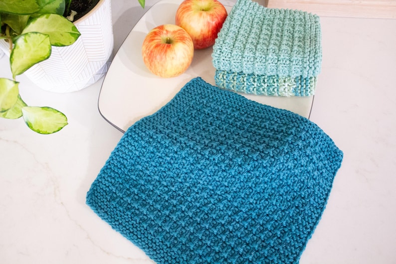 May include: A set of three square crochet washcloths featuring a textured waffle stitch pattern. One washcloth is a deep teal blue, while the other two are a lighter sage green. The cloths are arranged with two red and yellow apples nearby.