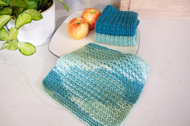 May include: Three square crocheted washcloths featuring a textured stitch pattern. One cloth displays a variegated gradient of teal, blue, and light green, while the other two are solid colors in dark teal and light seafoam green respectively.