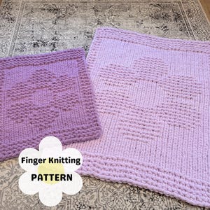 Könnte beinhalten: Zwei gestrickte quadratische Decken in Lilatönen, eine kleinere und eine größere, mit einem Herzmuster in der Mitte. Die Decken sind aus dickem Garn gefertigt und haben ein strukturiertes Aussehen. Der Text "Finger Knitting PATTERN" befindet sich auf dem Bild.