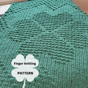 Op de afbeelding: Een grote, handgebreide deken in een teal groene kleur, met een klaverbladpatroon. De deken is gemaakt met dik garen, waardoor een textuur ontstaat. De woorden "Finger Knitting PATTERN" staan op een klaverblad.