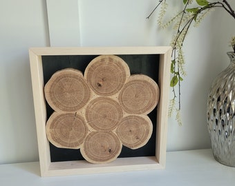 May include: A framed wooden art piece featuring seven circular wood slices arranged in a flower-like pattern against a black background. The frame is light-colored wood, and the piece is displayed on a white surface.