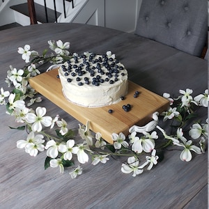 May include: A white cake with blueberries on top, sitting on a wooden serving board with a white handle. The board is surrounded by white flowers.