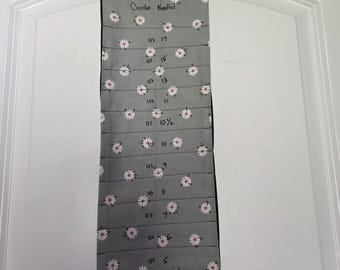 May include: A gray fabric organizer for circular knitting needles featuring a white daisy pattern. The organizer has vertical slots labeled with US needle sizes from 2 to 17, with the text Circular Needles printed at the top and two fabric hanging loops.