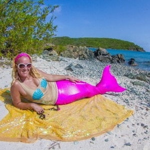 Pu&ograve; includere: Una donna con una coda di sirena rosa e un top blu e beige giace su una coperta di paillettes gialle su una spiaggia di sabbia bianca.