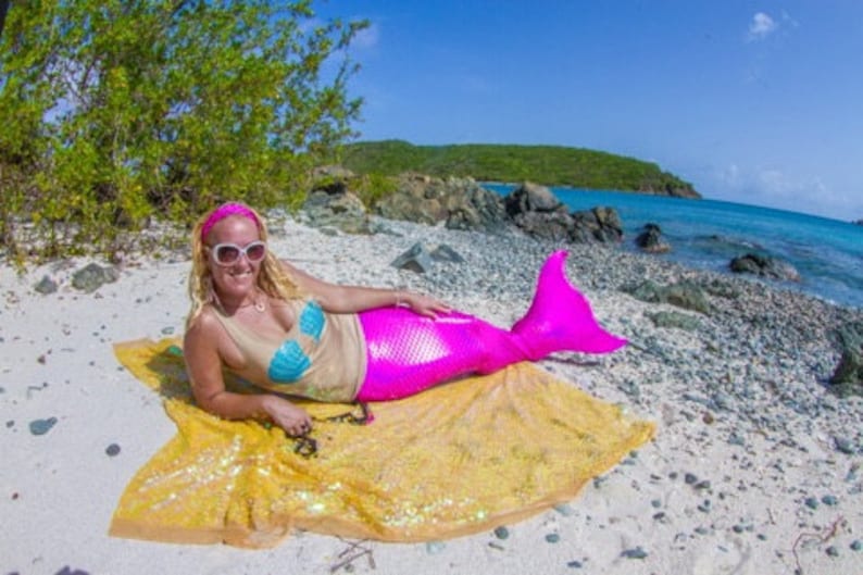 Pu&ograve; includere: Una donna con una coda di sirena rosa e un top blu e beige giace su una coperta di paillettes gialle su una spiaggia di sabbia bianca.