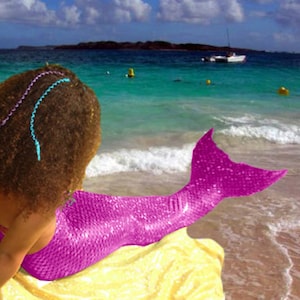 May include: A pink mermaid tail with a sparkly finish. The tail is worn by a person with curly brown hair on a beach.