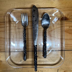 May include: A clear, rectangular glass plate holds a fork, knife, and spoon. The utensils have dark, twisted handles and are made of metal. The knife blade is long and the spoon is oval-shaped. The set is on a wooden surface.