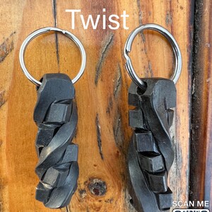 May include: Two metal keychains with a twisted cube design. The keychains are silver and black and have a ring at the top. The text "Cube Twist" is written above the keychains.