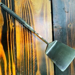 May include: A hand-forged metal spatula with a twisted handle and a rectangular head. The spatula is made of a dark, possibly blackened, metal. The handle has a hole for hanging. The spatula rests on a wooden surface.