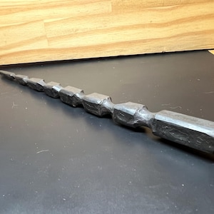 May include: A dark gray, hand-carved wooden tool with a faceted, geometric design. The tool tapers to a point on one end and has a rectangular handle on the other. The tool is approximately 10 inches long.