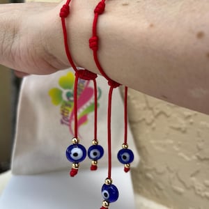 May include: Two red string bracelets featuring small blue evil eye beads and gold-toned metallic accents. Each bracelet has adjustable knots and dangling charms with a central blue eye design, white iris, and black pupil, finished with gold beads.