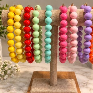 May include: A collection of eight beaded bracelets displayed on a T-shaped stand. The bracelets feature smooth, round, solid-colored beads in yellow, red, light green, turquoise, hot pink, light pink, purple, and orange, each with a gold-toned clasp.