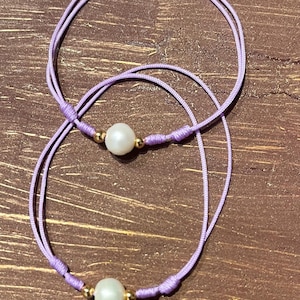 Puede incluir: Dos pulseras de hilo lavanda con una perla blanca y detalles de cuentas doradas. Las pulseras están anudadas y descansan sobre una superficie de madera. La joyería es delicada y minimalista.