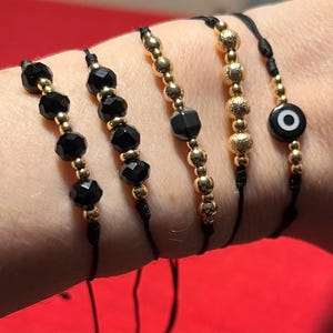 May include: Five black and gold beaded bracelets on black cords. The bracelets feature faceted black beads, gold-colored beads, and an evil eye charm. The bracelets are displayed against a red background.