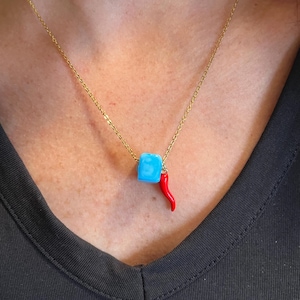 May include: A gold-colored chain necklace featuring a light blue square bead and a small red chili pepper charm. The necklace is worn against a dark background.