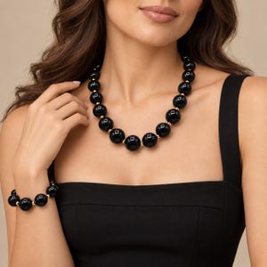May include: A necklace and bracelet set featuring large, glossy black spherical beads alternating with small, polished gold-tone metal spacer beads. The jewelry pieces are strung on a cord, creating a uniform, rounded design with a high-shine finish.