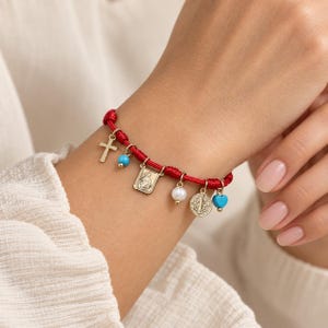 Peut inclure: Un bracelet en cordon rouge orné de plusieurs breloques dorées, dont une croix, un pendentif religieux rectangulaire, une médaille circulaire, une perle blanche, une petite perle turquoise et un cœur turquoise, fixés par des anneaux dorés.