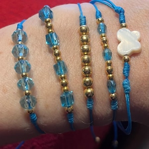 May include: Five blue cord bracelets with different bead arrangements. The bracelets have gold-coloured beads, clear blue crystal beads, and a white clover-shaped charm. The bracelets are shown on a wrist.