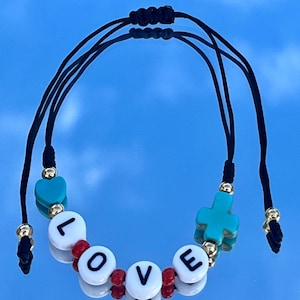 Può includere: Braccialetto regolabile con cordino nero con perline a lettera bianche che compongono "LOVE". Il braccialetto presenta ciondoli a forma di cuore e croce turchese, perline rosse e dettagli metallici dorati. Lo sfondo è una superficie blu riflettente.