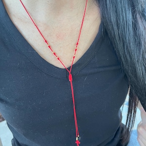 May include: Red string necklace with gold beads and a small red cross pendant. The necklace is worn against a black shirt. The Y-shaped design features a knotted detail and a dangling strand.