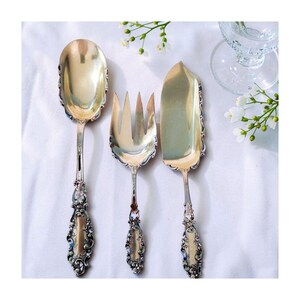 May include: A three-piece silver serving set consisting of a large spoon, a three-tined fork, and a flat server. Each piece features a highly polished metallic finish and an ornate, embossed handle with intricate scrollwork and floral detailing.