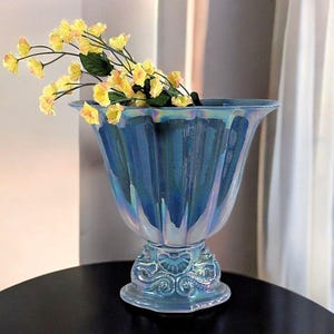 May include: A blue iridescent glass vase with a fluted design, holding yellow flowers. The vase has a decorative base with ornate detailing. The vase is sitting on a black surface.