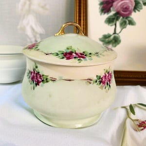 May include: A vintage ceramic jar with a lid, featuring hand-painted pink roses and green leaves on a cream-colored background. The lid has a gold handle. The jar is likely used for storing small items.