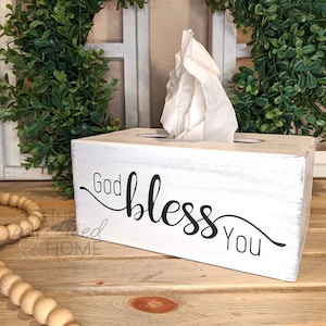 May include: A white wooden tissue box with the words "God bless You" in black script. The box is rectangular with a distressed finish, and a bundle of white tissues is visible. The box is on a wooden surface.
