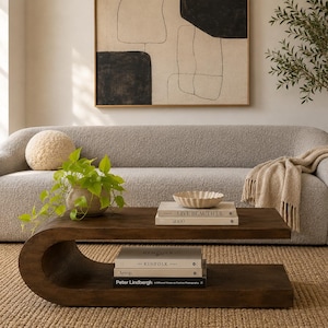 May include: A curved, dark wood coffee table with a lower shelf, featuring a stack of books titled Live Beautiful, Kinfolk, and Peter Lindbergh, topped with a small, fluted ceramic bowl and a potted green plant on the upper surface.