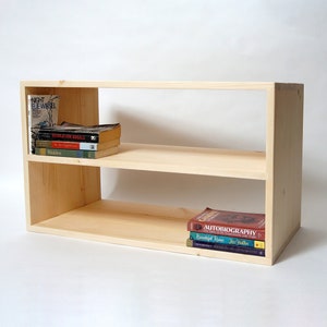 May include: A light-colored wooden bookshelf featuring two open horizontal shelves. The top shelf holds a stack of four books, including Night by Elie Wiesel and Walden. The bottom shelf holds a stack of three books, including an Autobiography.