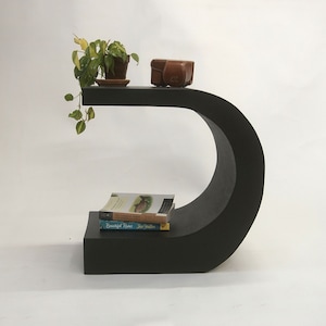 May include: A modern, black, sculptural side table with a curved design. The table has a top shelf with a small potted plant and a brown leather case. Two books are stacked on the lower shelf. The table is approximately 30 inches long.