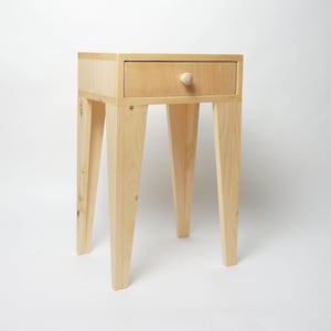 May include: A light-coloured wooden bedside table with a single drawer and a round knob. The table has four angled legs and is made of light-coloured wood. The drawer is centred on the front of the table.