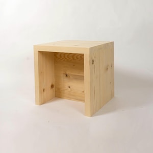 May include: A light-colored, unfinished wooden cube-shaped side table or stool. The open design features a square top and a hollow interior, with two vertical supports. The wood grain is visible, and the piece is set against a white background.
