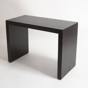 May include: A rectangular console table constructed from solid wood with a matte black finish. The piece features a continuous waterfall design where the flat top transitions seamlessly into two vertical side panels, showcasing a visible wood grain.