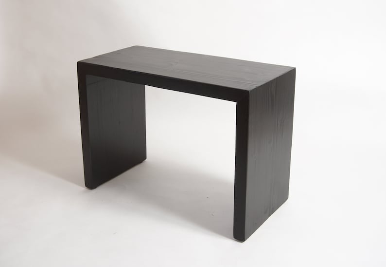 May include: A rectangular console table constructed from solid wood with a matte black finish. The piece features a continuous waterfall design where the flat top transitions seamlessly into two vertical side panels, showcasing a visible wood grain.