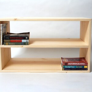 May include: A rectangular light-colored wooden bookshelf with two open shelves. The top shelf holds a vertical book and a stack of four books. The bottom shelf holds a stack of three books. The unit has a smooth, unfinished wood grain finish.