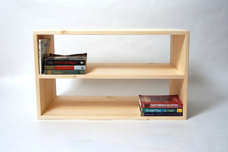 May include: A rectangular light-colored wooden bookshelf with two open shelves. The top shelf holds a vertical book and a stack of four books. The bottom shelf holds a stack of three books. The unit has a smooth, unfinished wood grain finish.