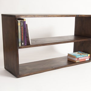 May include: A rectangular dark brown wooden bookshelf featuring two horizontal shelves. The top shelf contains four upright books on the left side, while the bottom shelf holds a small stack of three books positioned on the right side.