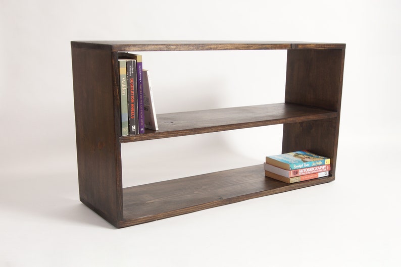 May include: A rectangular dark brown wooden bookshelf featuring two horizontal shelves. The top shelf contains four upright books on the left side, while the bottom shelf holds a small stack of three books positioned on the right side.