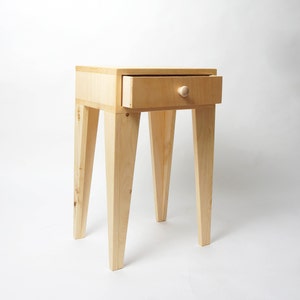May include: A small, unfinished wooden side table with a single drawer. The drawer is open, revealing the interior. The table has four angled legs and a round wooden knob. The table is approximately 61 cm tall.