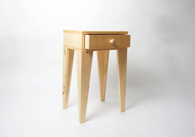 May include: A small, unfinished wooden side table with a single drawer. The drawer is open, revealing the interior. The table has four angled legs and a round wooden knob. The table is approximately 61 cm tall.
