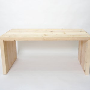 May include: A rectangular table constructed from light-colored natural wood with visible grain and knots. The design features a flat top surface supported by two vertical side panels that connect to the top at mitered corner joints.