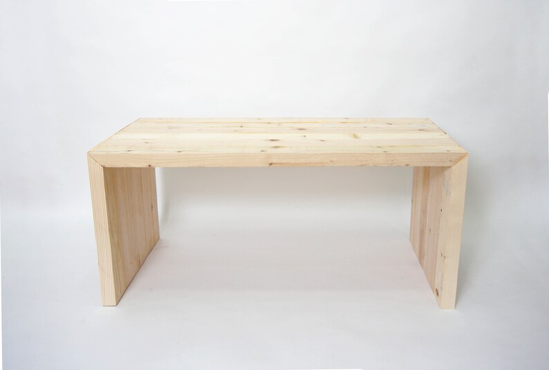 May include: A rectangular table constructed from light-colored natural wood with visible grain and knots. The design features a flat top surface supported by two vertical side panels that connect to the top at mitered corner joints.
