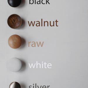 May include: Five round cabinet knobs arranged vertically. The top knob is black, followed by a brown walnut wood knob, a light tan raw wood knob, a matte white knob, and a metallic silver knob. Each knob is labeled with its color name in text.