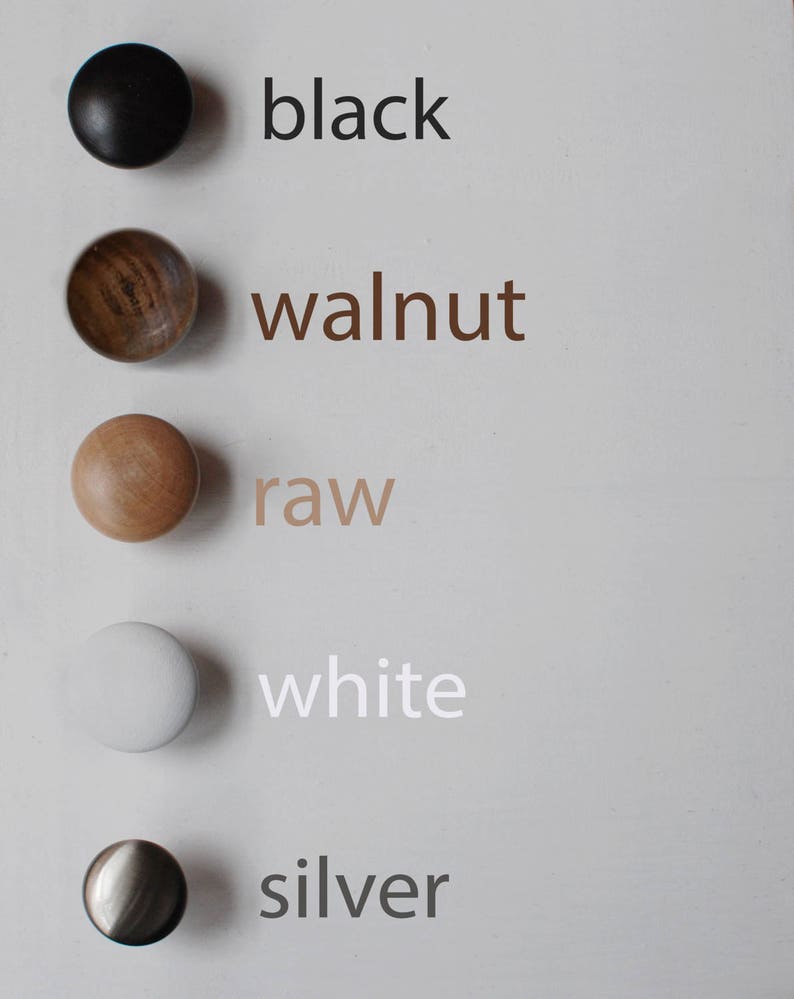 May include: Five round cabinet knobs arranged vertically. The top knob is black, followed by a brown walnut wood knob, a light tan raw wood knob, a matte white knob, and a metallic silver knob. Each knob is labeled with its color name in text.