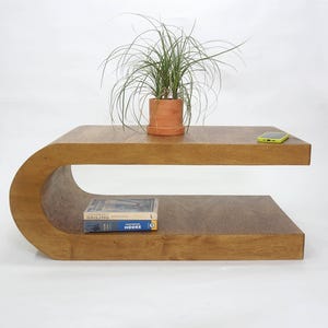 May include: A C-shaped wooden coffee table with a smooth brown finish. A terracotta-colored pot containing a green grass-like plant sits on the top surface. A lime green smartphone and a book titled Sailing Moore are placed on the table.