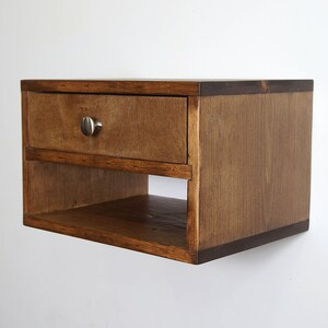 May include: A wall-mounted wooden floating nightstand featuring a rectangular frame with a medium brown wood grain finish. The unit includes a single top drawer with a small, round, brushed silver metal knob and an open storage shelf below.