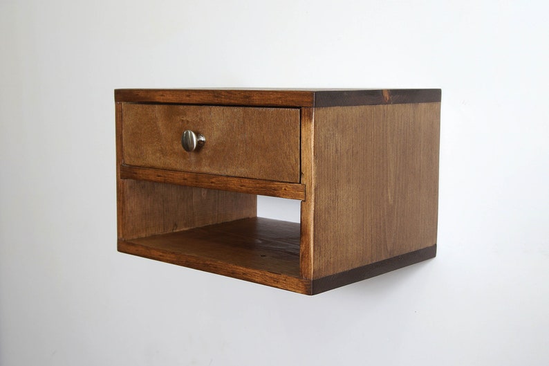 May include: A wall-mounted wooden floating nightstand featuring a rectangular frame with a medium brown wood grain finish. The unit includes a single top drawer with a small, round, brushed silver metal knob and an open storage shelf below.