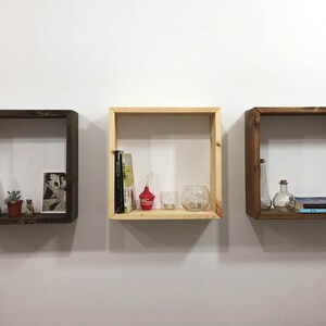 May include: Three square wooden wall shelves mounted in a horizontal row. The two outer shelves are finished in a dark brown stain, while the center shelf is a natural light wood tone. Each shelf contains various books, small plants, and glass items.
